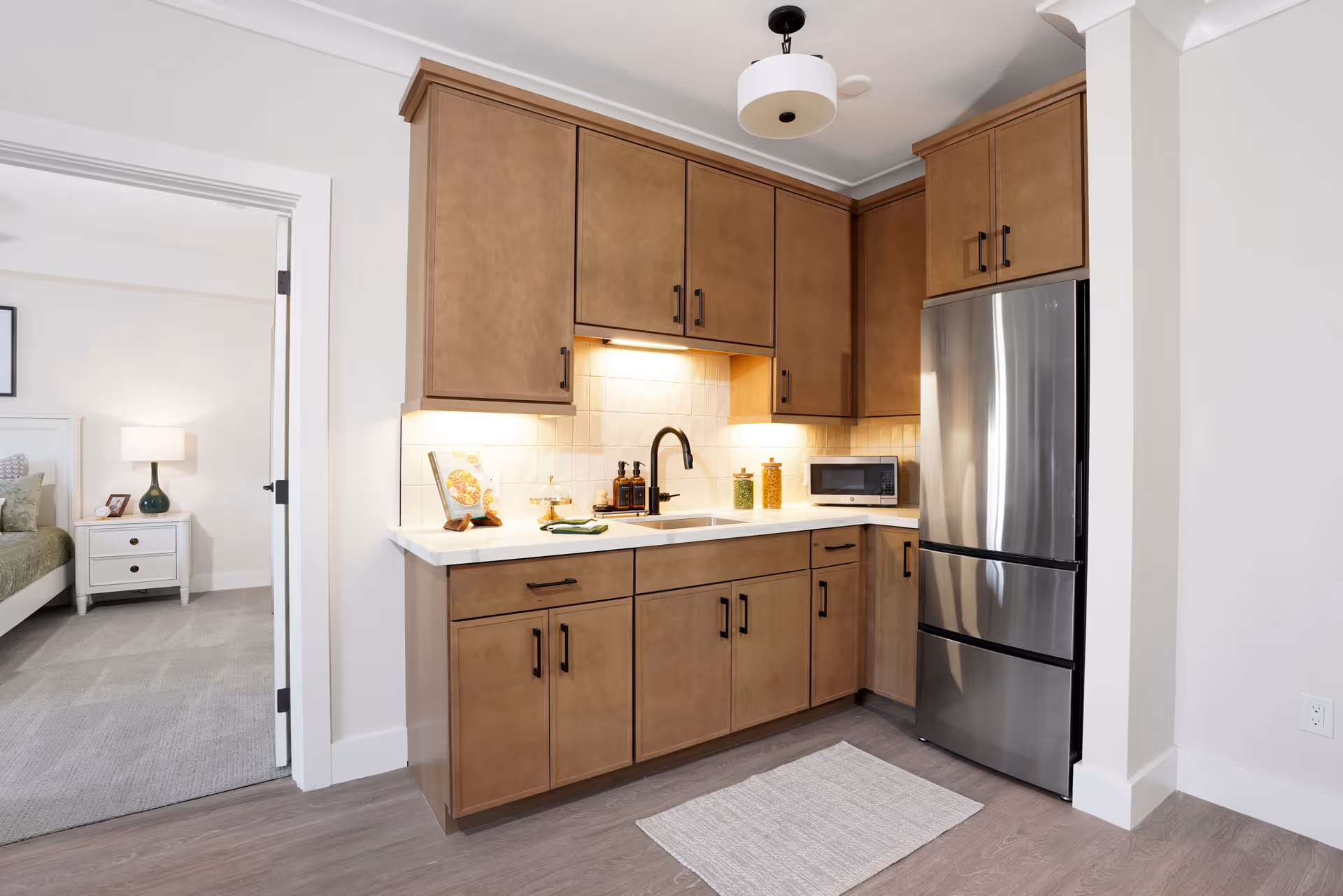 Small modern kitchen with wood cabinets, a stainless steel refrigerator, sink, and microwave, with a bedroom visible through a doorway.