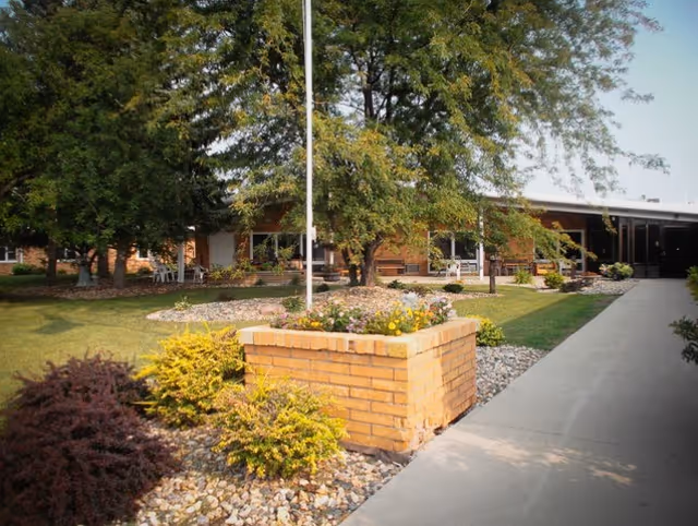 Outdoor view of a senior living facility with a brick planter filled with flowers in the foreground, surrounded by small bushes and rocks. A concrete walkway leads to the building entrance, which is partially shaded by large trees.