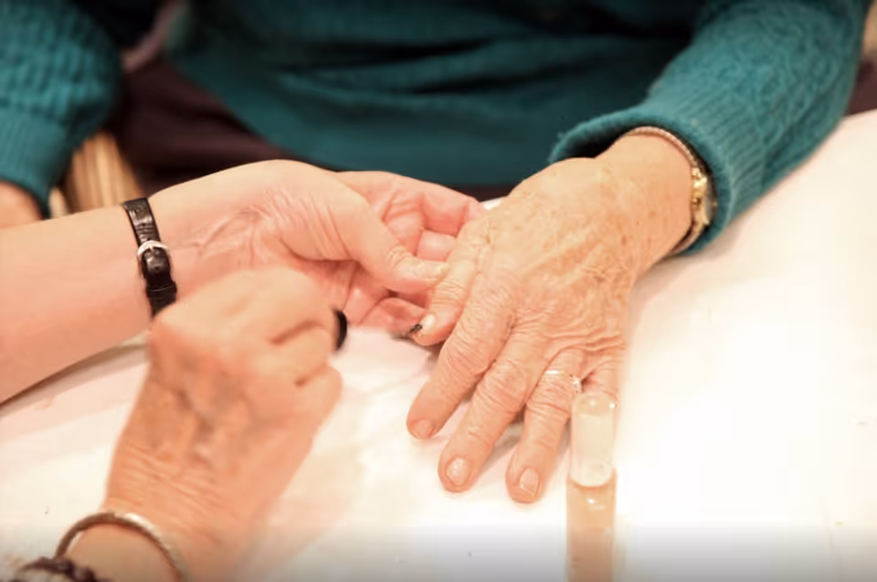 Close-up of two elderly hands, one person painting the fingernails of the other with clear nail polish on a white surface.