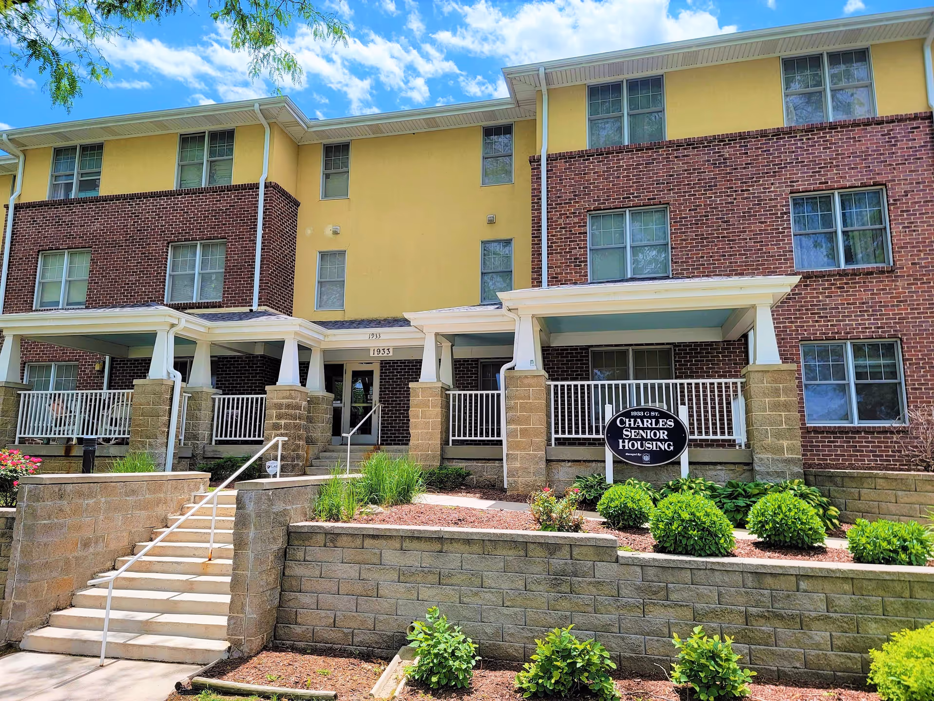 Exterior view of a three-story senior housing building with a combination of yellow and brick facade. There are stairs leading up to the entrance, white railings on the porches, and well-maintained landscaping with bushes and flowers. A sign in front reads '1933 G St Charles Senior Housing'.