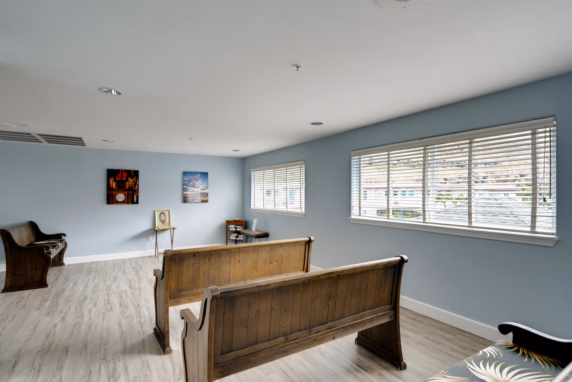 A quiet room with light blue walls and wood flooring featuring wooden pew-style benches arranged facing a wall with two framed pictures and a small table with a framed photo. Large windows with white blinds allow natural light to enter the room.
