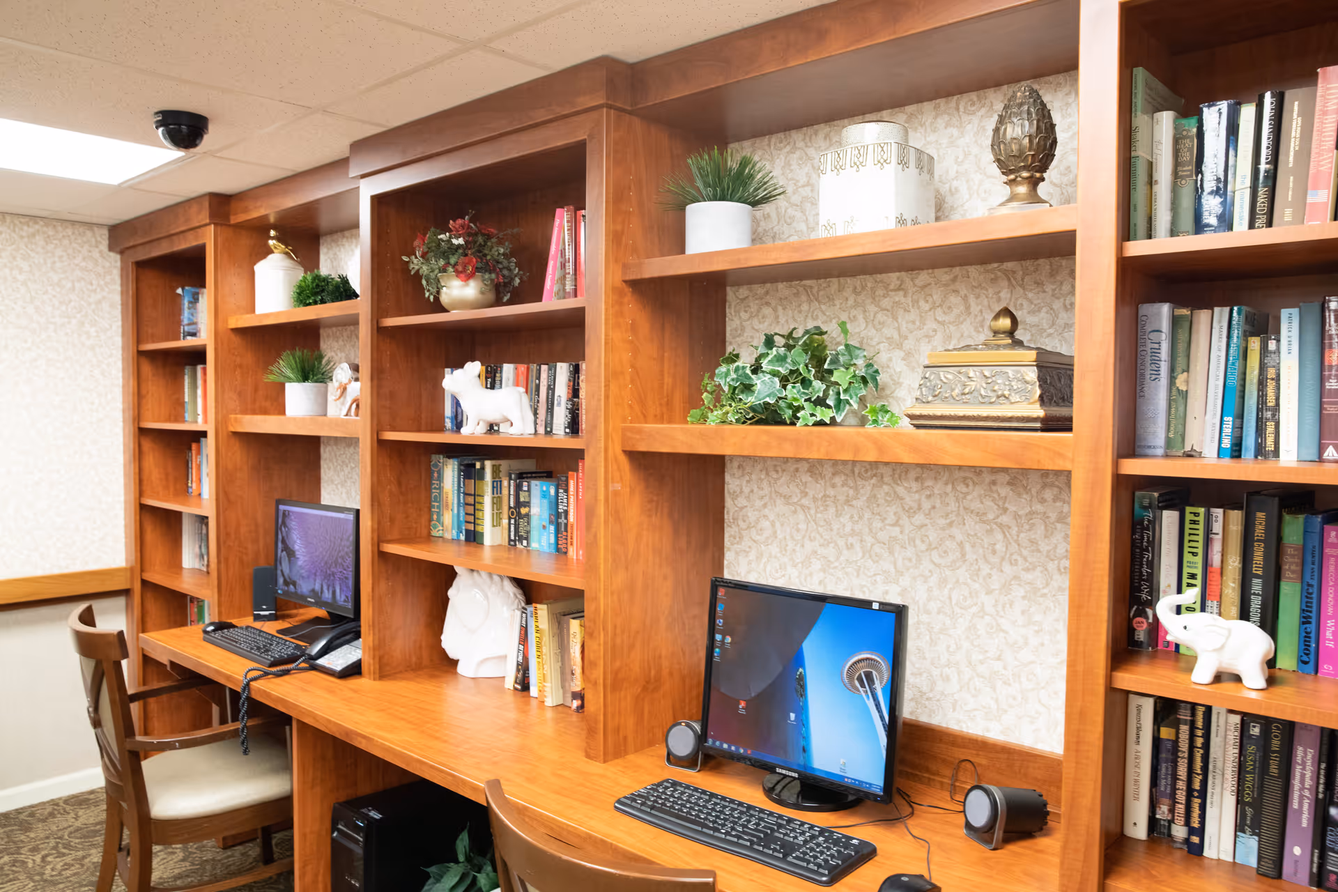 A cozy computer workstation area in a senior living facility with wooden built-in bookshelves filled with books, decorative plants, and ornaments. Two desktop computers with keyboards and speakers are placed on a wooden desk with chairs. The background features patterned wallpaper and a ceiling with recessed lighting.