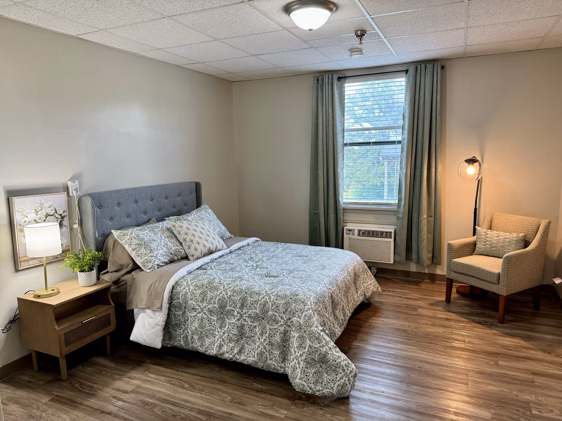 A cozy bedroom with a gray upholstered bed featuring patterned bedding and multiple pillows. Next to the bed is a wooden nightstand with a lamp, a small plant, and a framed floral picture on the wall. A window with green curtains and blinds is centered on the far wall, with an air conditioning unit below it. To the right, there is a beige armchair with a decorative pillow and a floor lamp with a round glass shade. The room has wood flooring and neutral-colored walls.