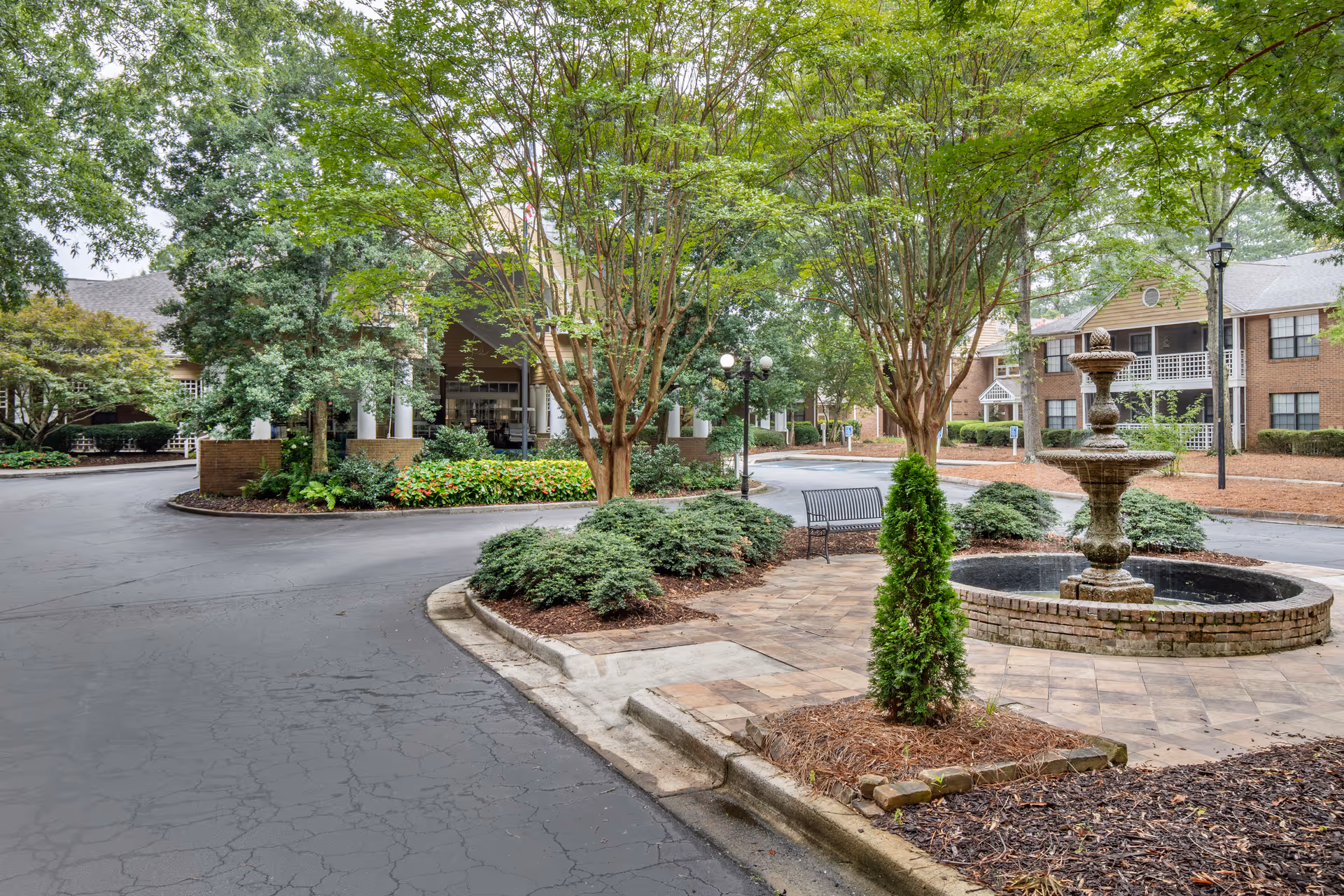 Outdoor view of a senior living facility with a circular driveway, landscaped greenery, trees, a stone fountain, a bench, and brick buildings in the background.