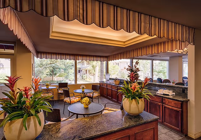 Interior view of a common area in a senior living facility with round tables and chairs, large windows showing trees outside, decorative plants in large pots on a granite countertop, and a striped valance around a recessed ceiling light.