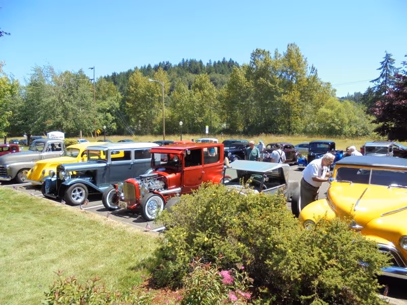 A parking lot filled with vintage and classic cars on a sunny day. Several people are walking around and looking at the cars. The background shows green trees and a clear blue sky. There is a grassy area with bushes and flowers in the foreground.