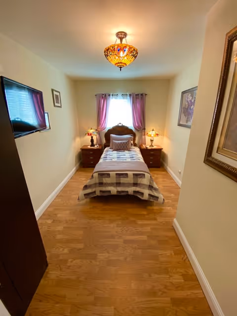A small, neatly arranged bedroom with a single bed centered against the back wall. The bed has a patterned quilt and a decorative pillow. On either side of the bed are wooden nightstands with matching Tiffany-style lamps. A window with purple curtains is above the bed, and a flat-screen TV is mounted on the left wall. The room has wooden flooring and light-colored walls with framed artwork.