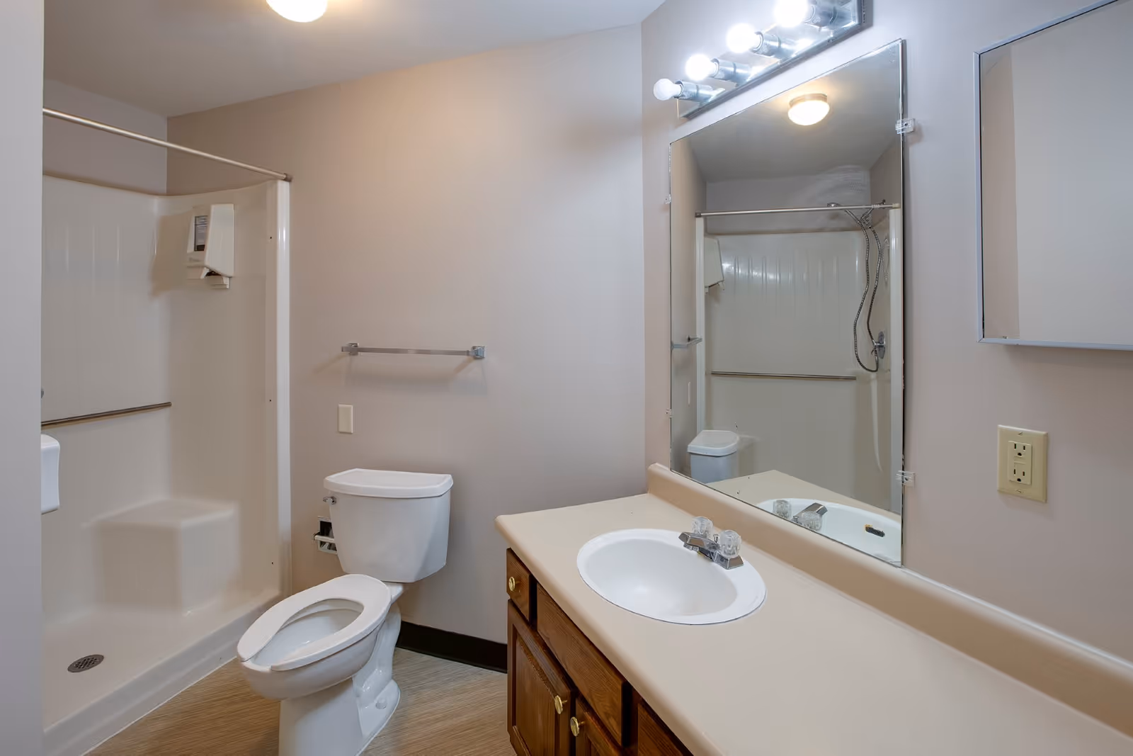 Bathroom with a toilet, sink and wooden vanity, large mirror, and a walk-in shower.