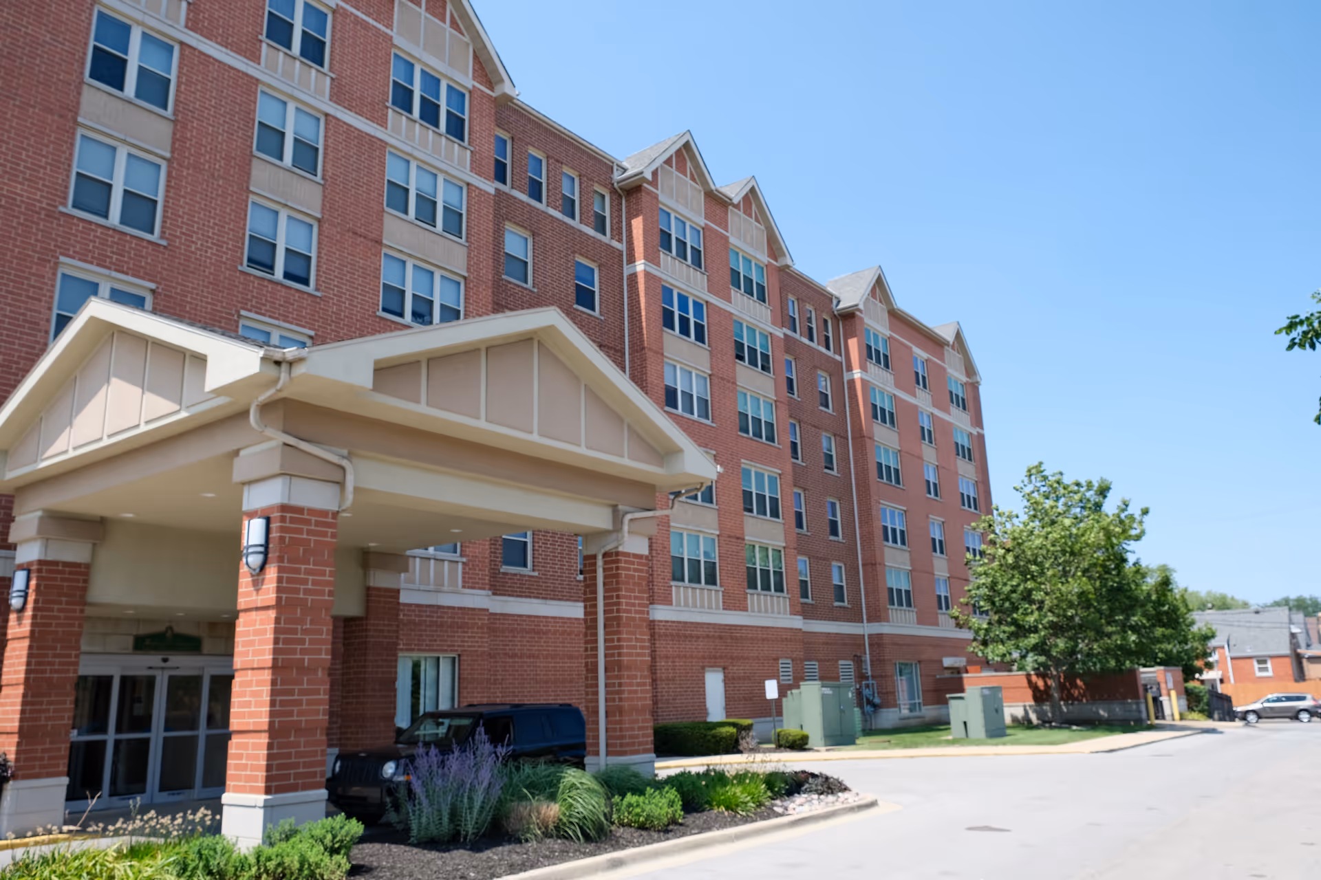 Brick multi-story senior living building with a covered entrance and driveway under a clear blue sky.