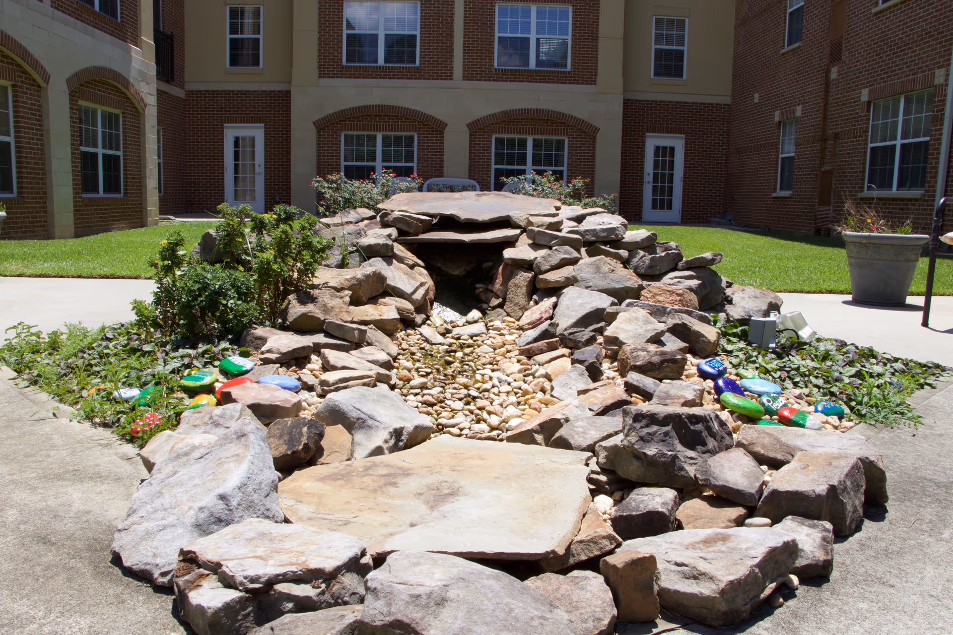A landscaped courtyard area with a dry rock stream bed surrounded by large stones and greenery. The courtyard is enclosed by a multi-story brick building with multiple windows and doors. There are painted rocks placed along the sides of the rock stream bed.