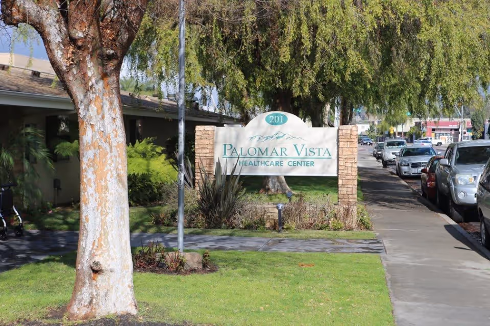 Entrance sign reading "Palomar Vista Healthcare Center" by a sidewalk with trees, grass, and parked cars.