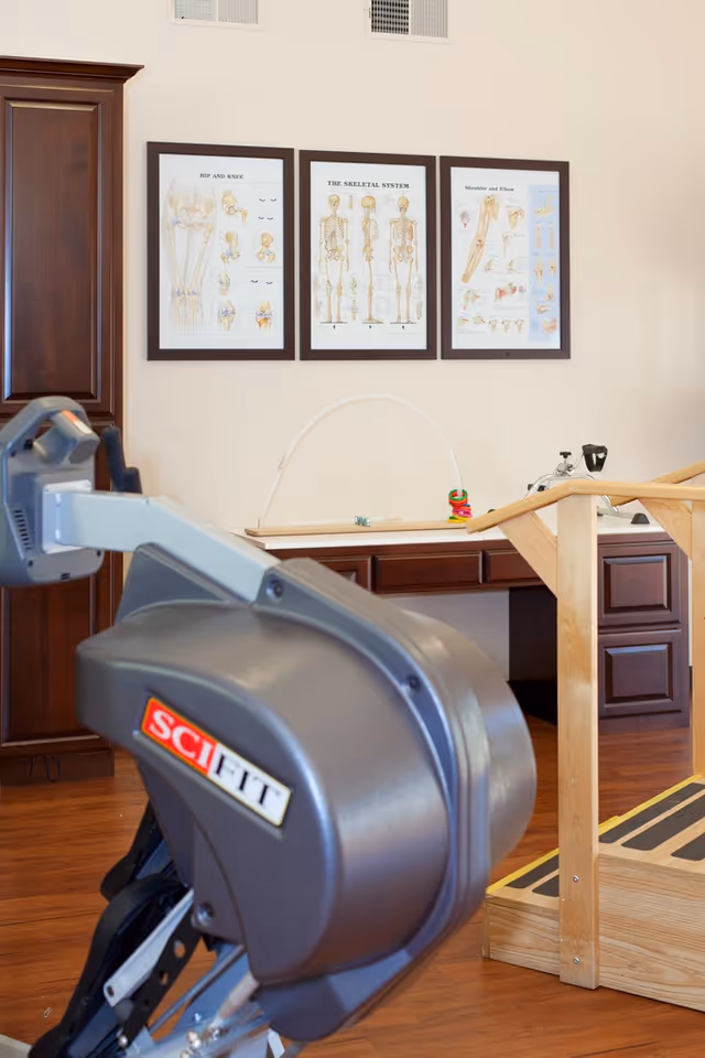 Physical therapy room with exercise equipment including a SCI FIT machine and wooden parallel bars. Three framed anatomical charts of the hip and knee, the skeletal system, and shoulder and elbow are displayed on the wall above a wooden cabinet.