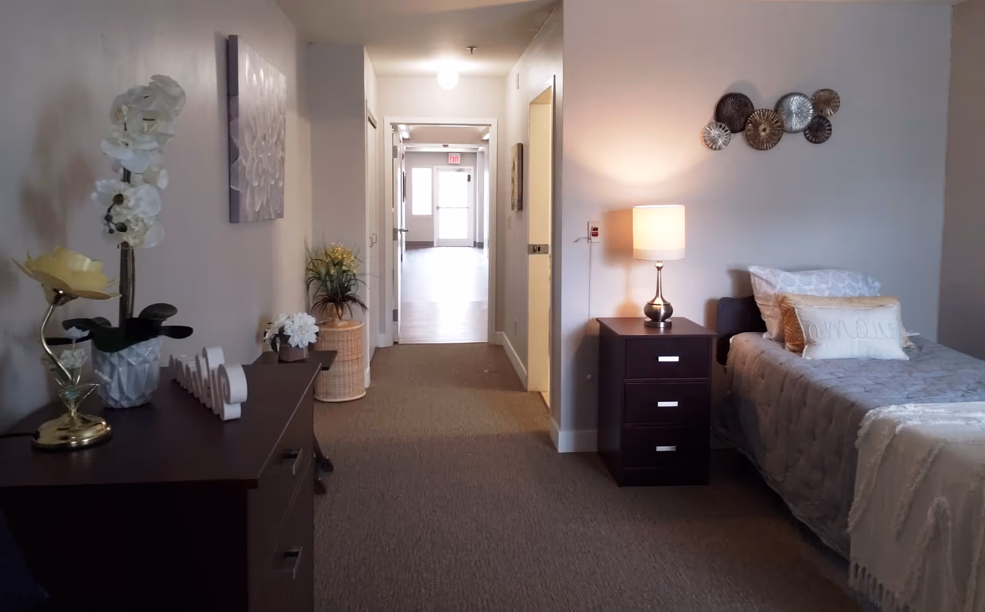 A cozy bedroom in an assisted living facility featuring a single bed with gray bedding and decorative pillows, a dark wooden nightstand with a lamp, and a dresser with decorative items including a white orchid and a yellow flower. The room has neutral-colored walls and carpeted floor, with a hallway leading to a door at the far end.