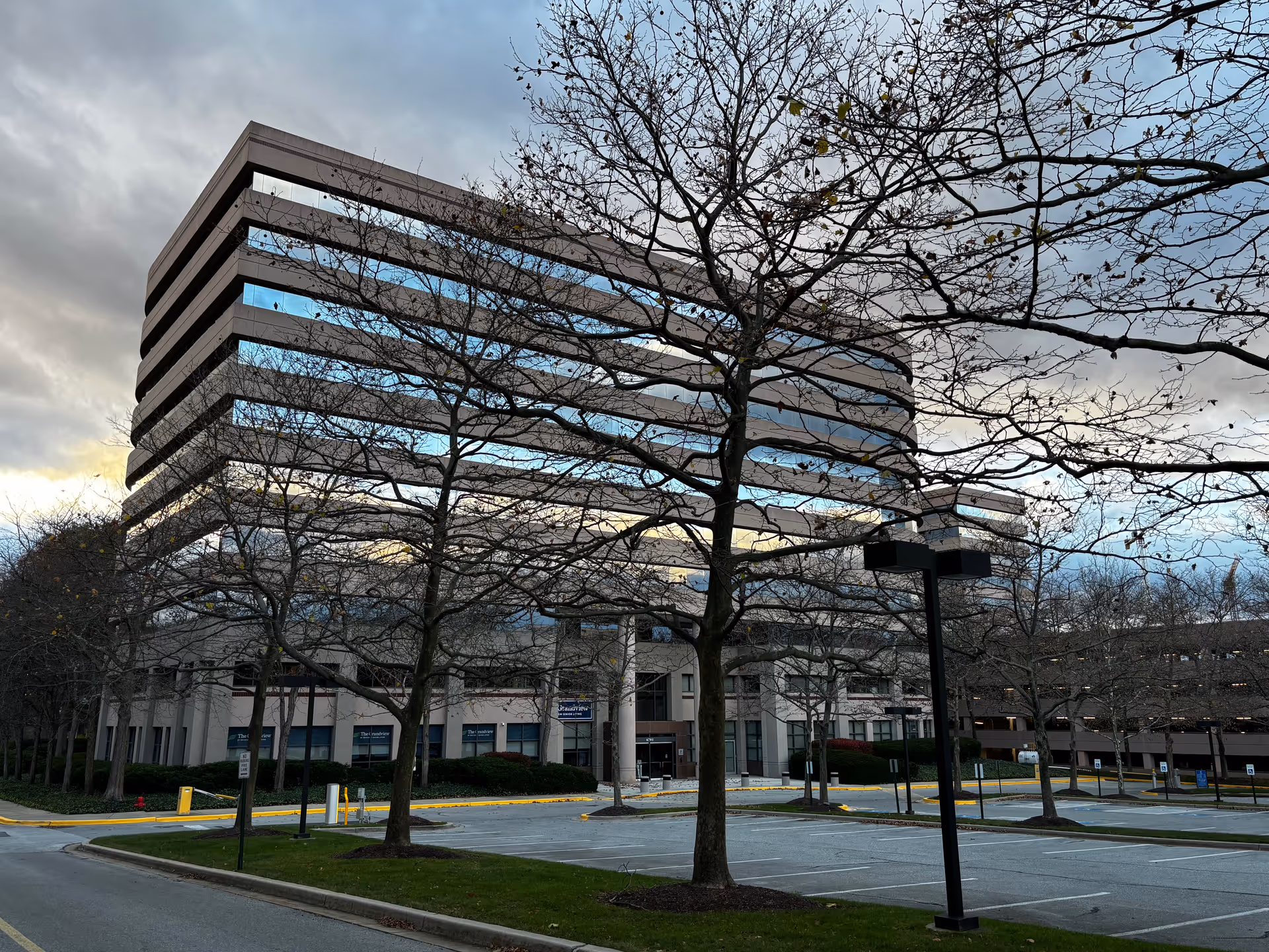 A large multi-story office building with reflective windows, surrounded by leafless trees and an empty parking lot under a cloudy sky.