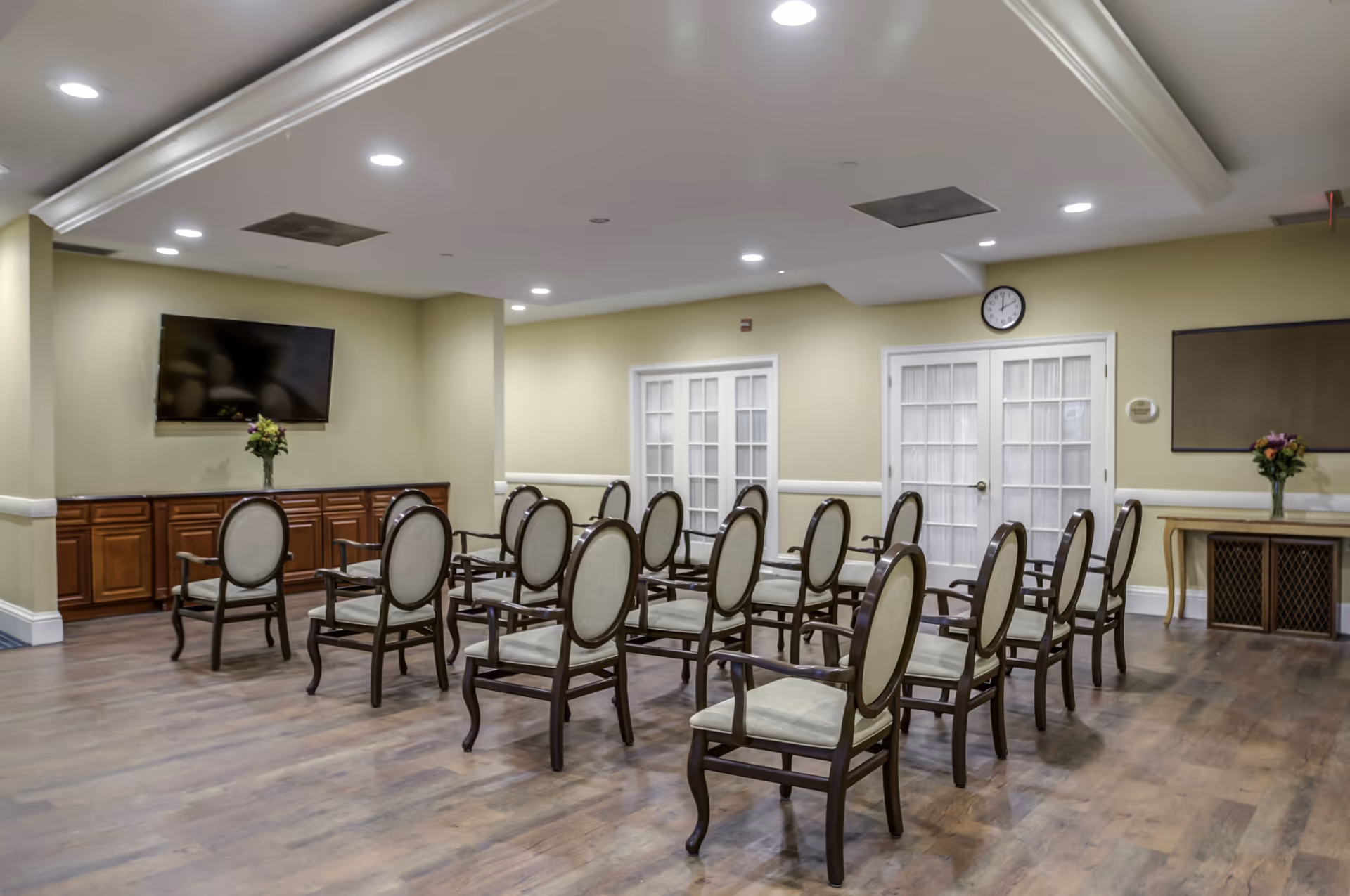 A meeting or activity room with multiple wooden chairs arranged in rows facing a wall-mounted flat screen TV. The room has light yellow walls, wooden flooring, recessed ceiling lights, a clock above double glass doors, and flower vases on a cabinet and a table.