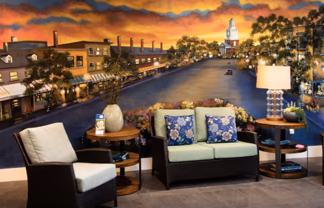 A cozy seating area with a cushioned armchair and a loveseat with floral pillows, flanked by two round wooden side tables. One table has a vase with flowers and the other has a lamp and small decorative items. Behind the seating area is a large mural depicting a scenic town street at sunset with buildings, trees, and a church steeple in the background.