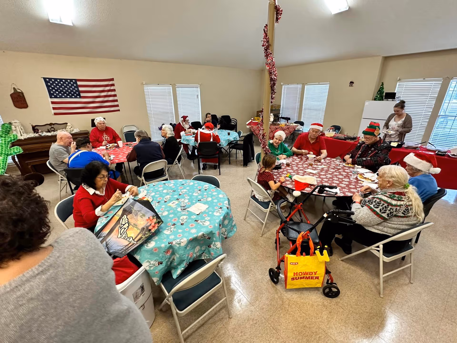 Seniors in holiday attire seated around decorated tables in a communal dining/activity room.