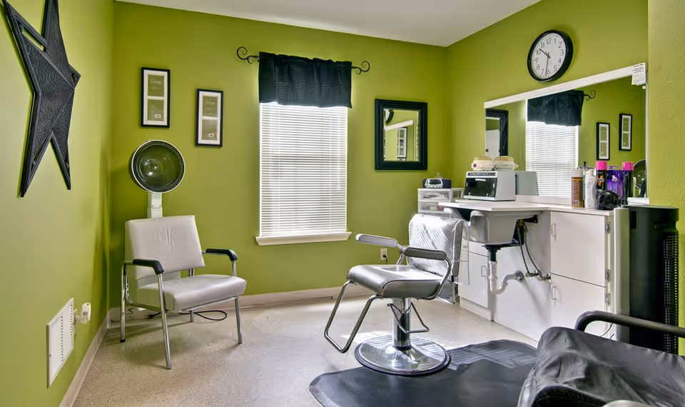 A small salon room with bright green walls featuring a salon chair, a white chair, a hair dryer, a large mirror above a white counter with various hair care products, and a clock on the wall showing 12:15.