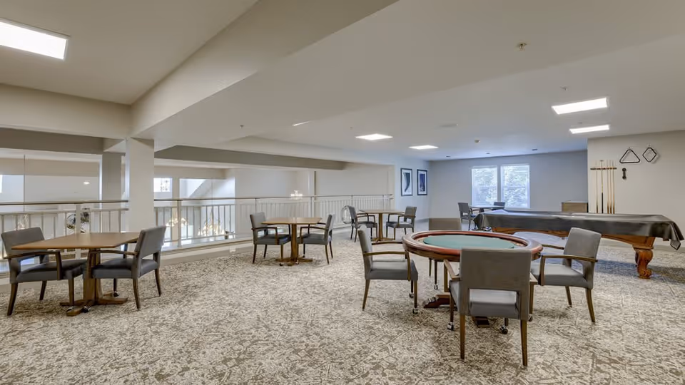 A spacious common area with multiple round tables and chairs arranged for seating. In the background, there is a covered pool table with cues and racks mounted on the wall. The room has large windows letting in natural light and neutral-colored walls and carpet.