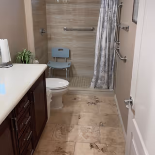 A bathroom with beige tiled floor and walls, featuring a white toilet, a dark wood vanity with a white countertop, a small green plant, and a walk-in shower with a blue shower chair and a patterned shower curtain. There are grab bars installed inside the shower and on the wall next to the door.