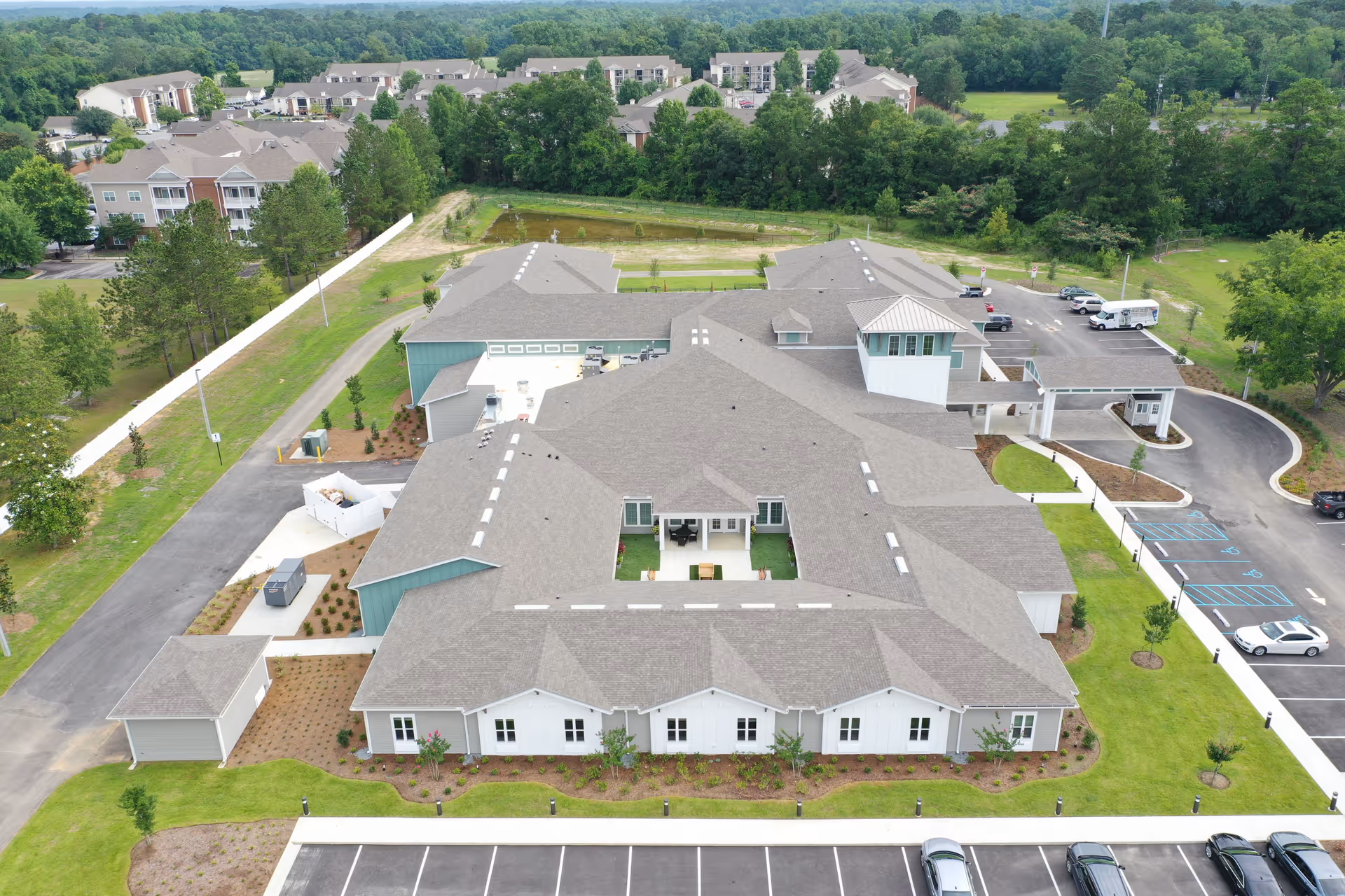 Aerial view of The Canopy at Hickory Creek senior living facility showing a large, modern building with multiple roof sections, surrounded by green lawns, parking lots, and trees. The building has a central courtyard with outdoor seating and is situated near other residential buildings and wooded areas.