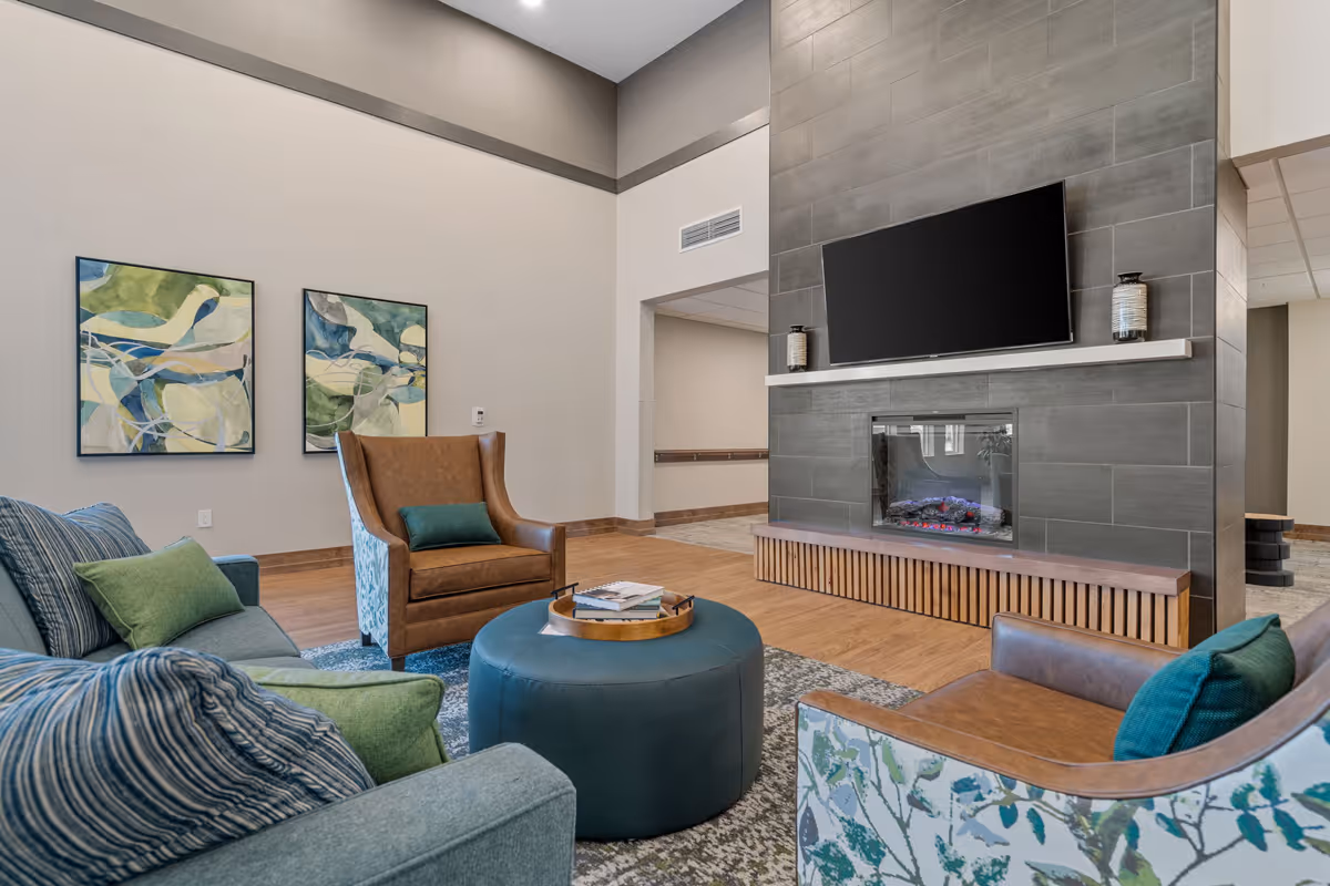 A cozy living room area in a senior living facility featuring a modern fireplace with a mounted flat-screen TV above it. The room has a mix of seating including a blue sofa with green and blue pillows, a brown leather armchair, and a floral-patterned chair. Two abstract paintings hang on the wall, and a round blue ottoman with a tray on top sits in the center on a patterned rug. The space has wooden flooring and neutral-colored walls.