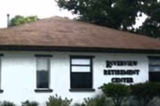 Exterior view of a single-story building with a brown roof and white walls. The building has three windows visible and a sign that reads 'Riverview Retirement Center'. There are some bushes and trees in front of the building.