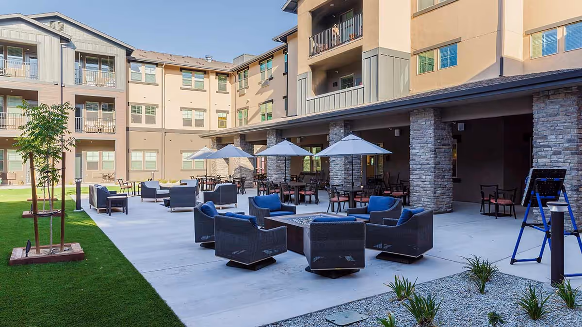 Outdoor patio area at Hilltop Springs Senior Living featuring multiple seating arrangements with cushioned chairs around a fire pit, tables with umbrellas, and a well-maintained lawn with young trees and landscaping. The patio is adjacent to a multi-story building with balconies and large windows under a clear blue sky.