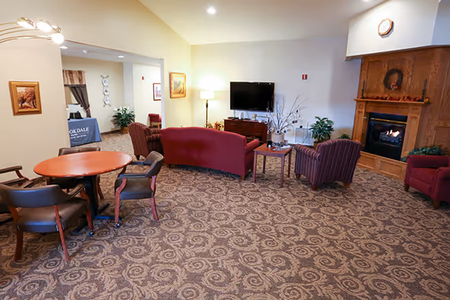 A cozy living room area in a senior living facility with patterned carpet, a round wooden table with four chairs, a curved red sofa, two striped armchairs, a wooden side table with a decorative vase, a flat-screen TV mounted on the wall, and a wooden fireplace with a wreath above it. The room is softly lit with a floor lamp and recessed ceiling lights.
