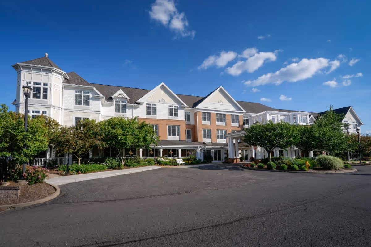 Front exterior of a three-story senior living building with a circular driveway, landscaped entrance, and blue sky.