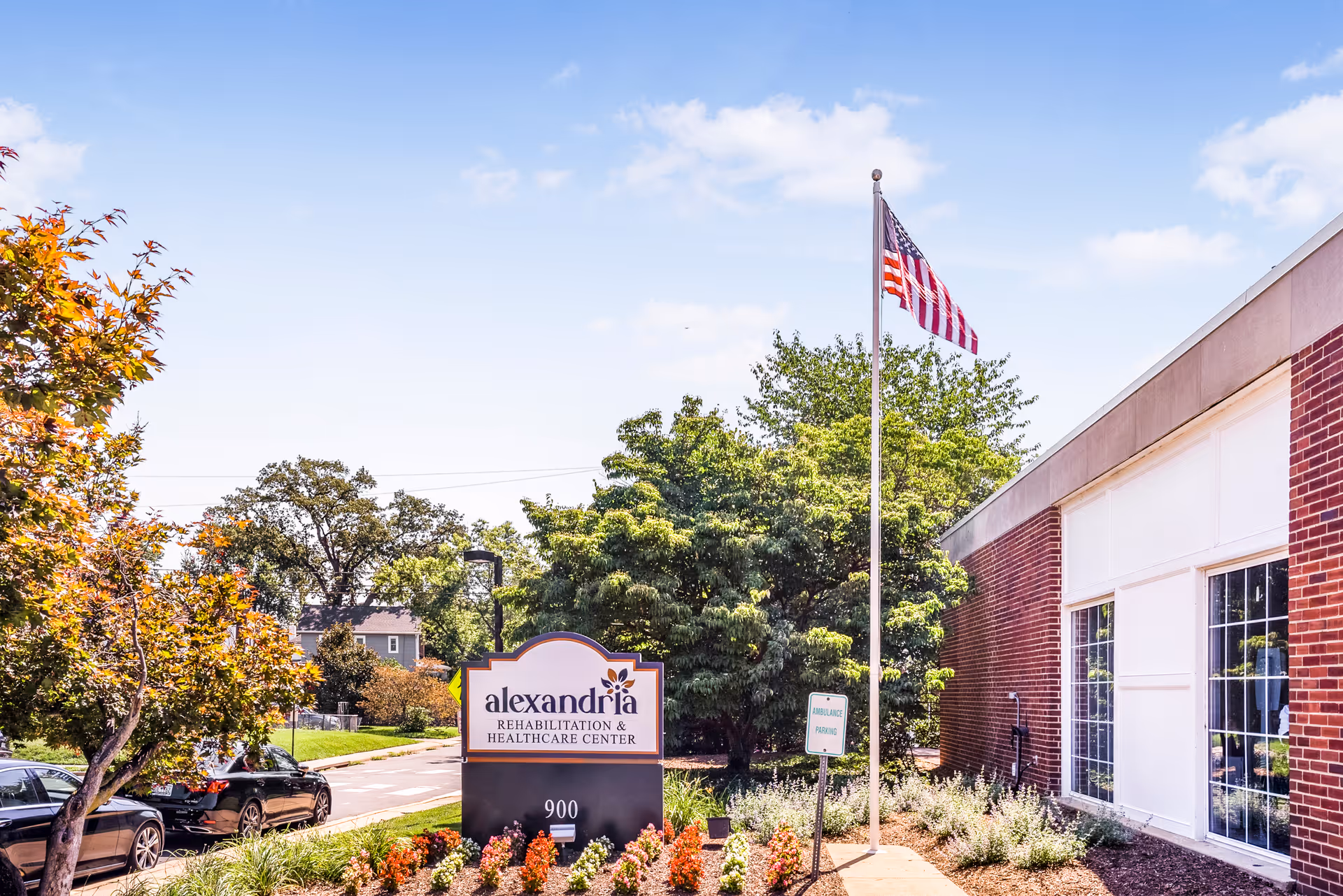 Exterior view of Alexandria Rehabilitation & Healthcare Center with a brick building on the right, an American flag on a flagpole, a sign displaying the facility's name, and landscaped plants and trees under a blue sky.