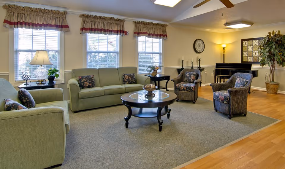 A cozy senior living facility common area with two green sofas, two wicker armchairs with floral cushions, a round glass-top coffee table, and side tables with lamps and plants. The room has three large windows with valances, a wall clock, a floor lamp, a piano, and a large potted plant in the corner. The floor is a combination of carpet and wood.