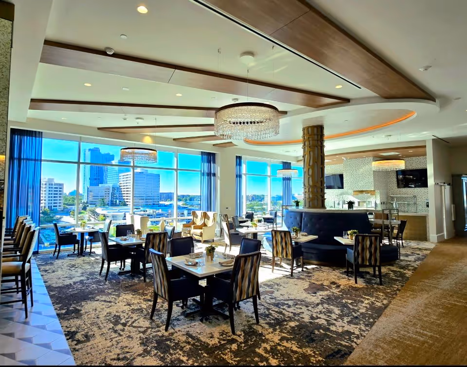 Bright and spacious dining area with multiple tables and chairs arranged neatly. Large windows provide a panoramic view of city buildings outside. The ceiling features wooden beams and elegant chandeliers. A circular booth seating area surrounds a decorative column in the center. The room has a modern and inviting atmosphere with patterned carpet and blue curtains.