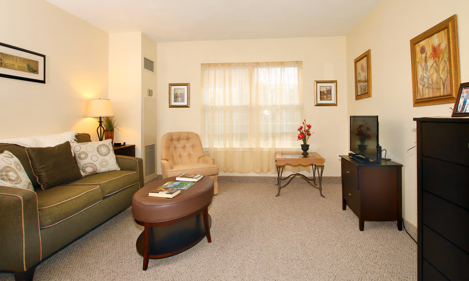 A cozy living room with a green sofa adorned with patterned cushions, a beige armchair, an oval coffee table with books on it, a side table with a lamp and plant, a small table with a vase of red flowers in front of a window with sheer curtains, a flat-screen TV on a dark wooden stand, and framed artwork on the walls.