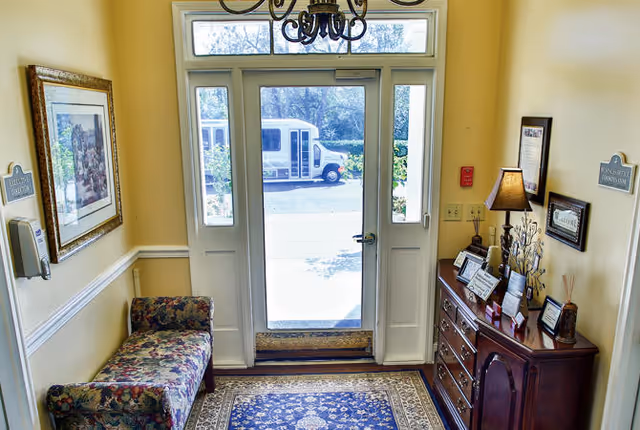 Entrance area of Dublin Square facility with a glass door and sidelights letting in natural light. A floral upholstered bench is on the left side under a framed picture, and a wooden dresser with a lamp, framed photos, and decorative items is on the right. A patterned rug covers the floor, and a chandelier hangs from the ceiling. Outside, a white shuttle bus is visible through the glass door.