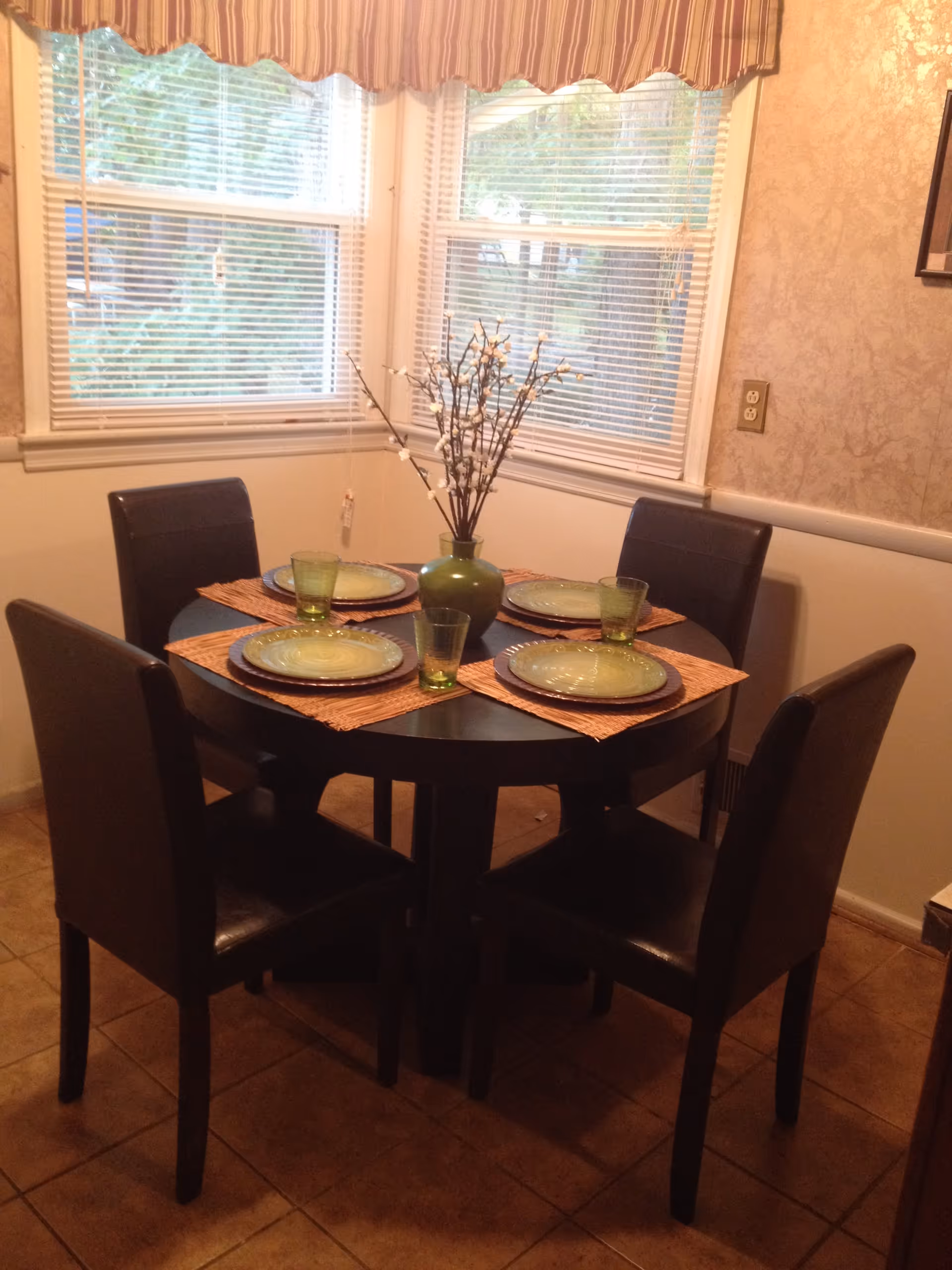 A dining area with a round dark wooden table set for four people. Each place setting has a green plate and a matching green glass on a woven placemat. In the center of the table is a green vase with decorative branches. The room has tiled flooring, beige walls with a textured pattern, and two windows with white blinds and a striped valance.