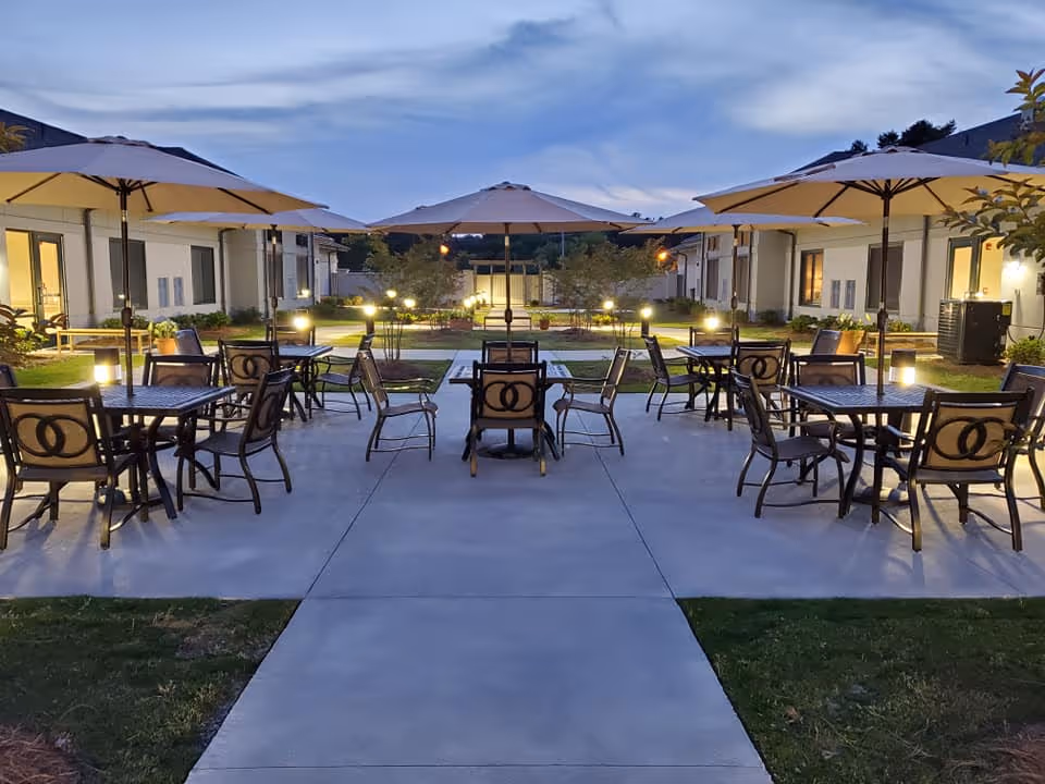 Evening courtyard patio with multiple tables, chairs and umbrellas arranged along a central walkway between single-story buildings with pathway lights.