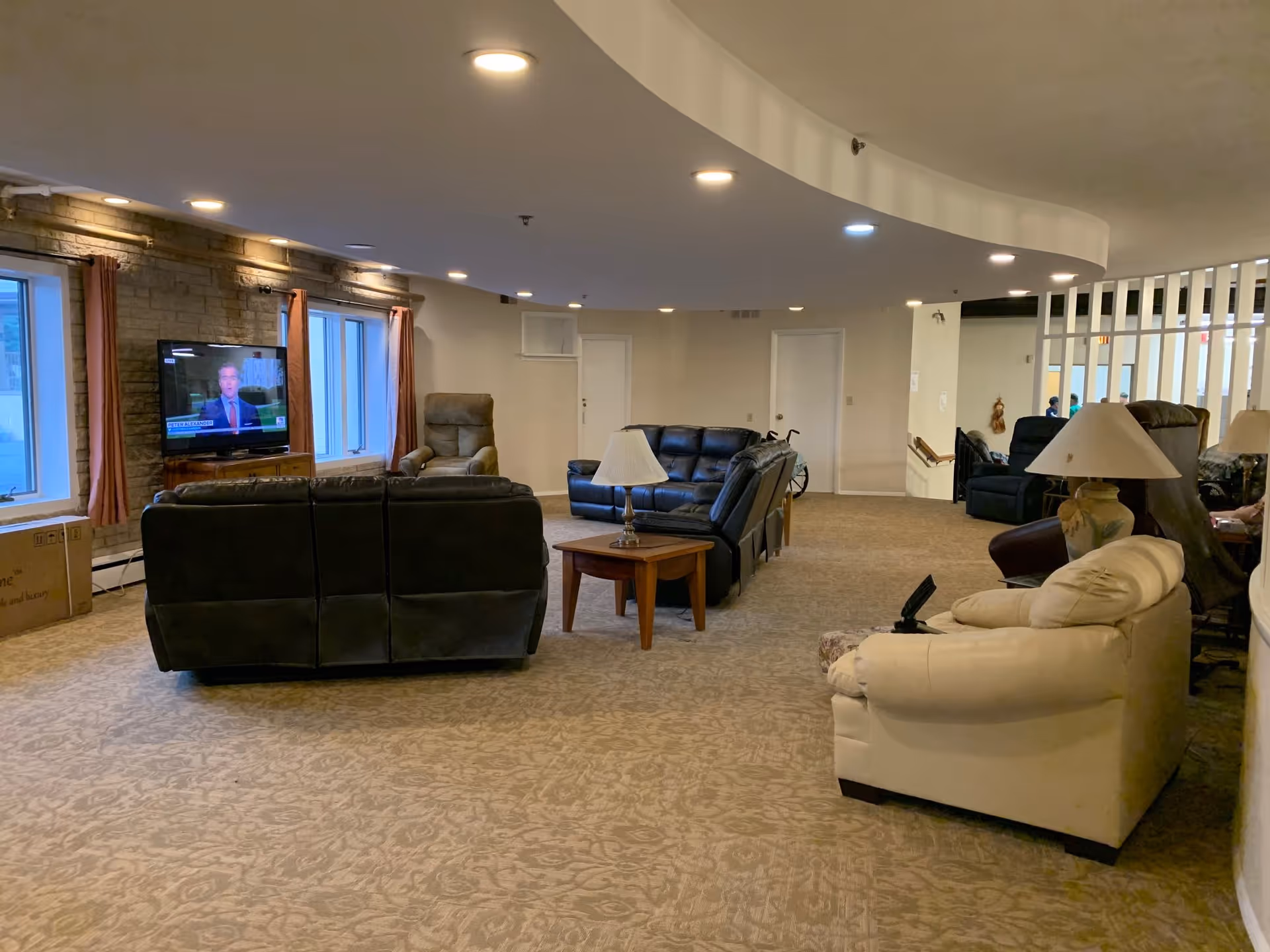 A spacious senior living common area with multiple leather sofas and armchairs arranged around wooden side tables with lamps. A flat-screen TV is mounted on a stone wall near two windows with curtains. The ceiling has recessed lighting, and the carpet has a subtle floral pattern. There is a wheelchair visible near the back wall.