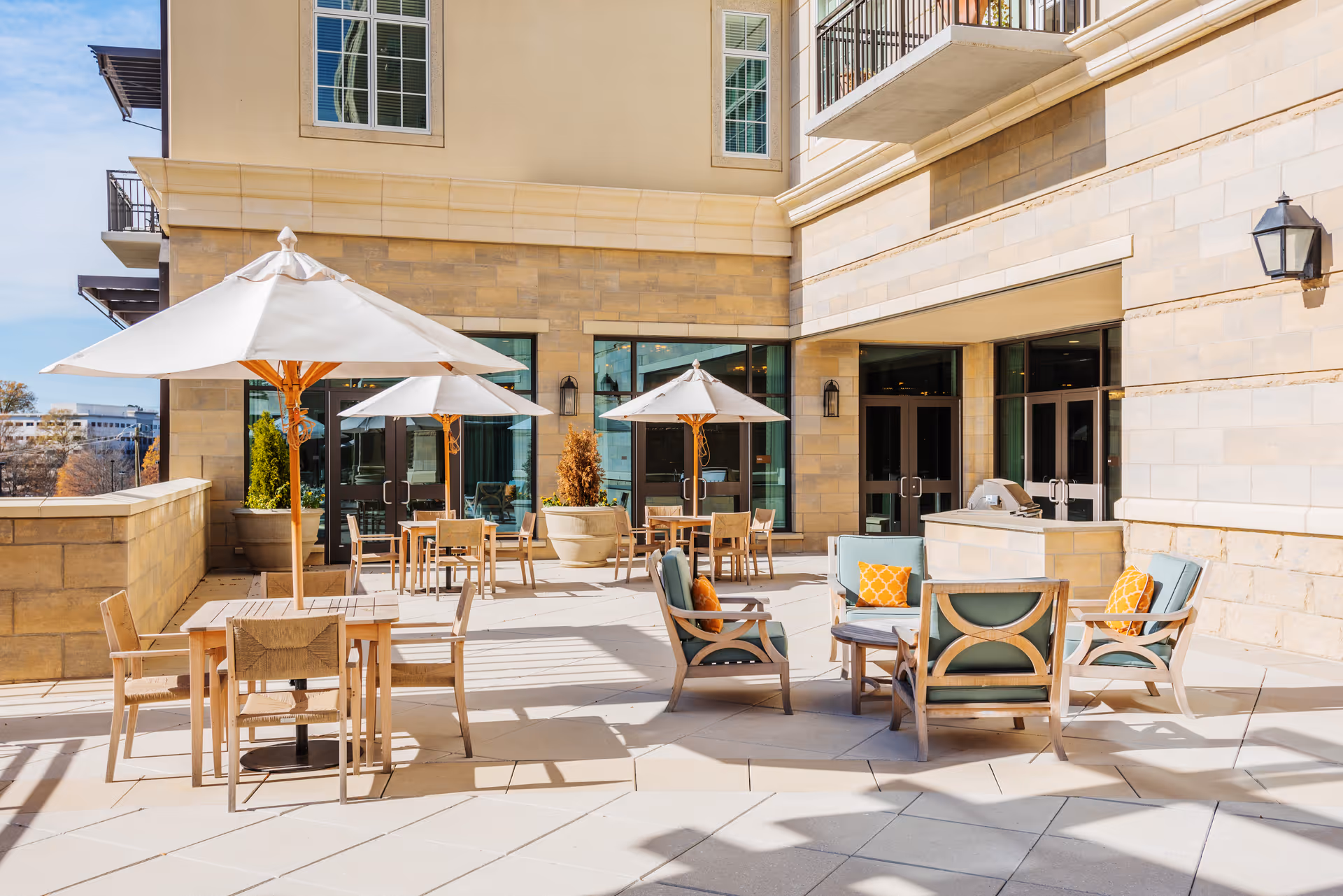Sunny outdoor patio with tables, umbrellas, and lounge seating in front of a stone-faced building.