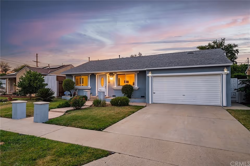 Exterior view of a single-story house with a gray roof and blue-gray walls at dusk. The house has a two-car garage with a white door, a front door with an oval window, and two lit windows. The front yard is landscaped with grass, bushes, and a concrete driveway and walkway leading to the entrance.