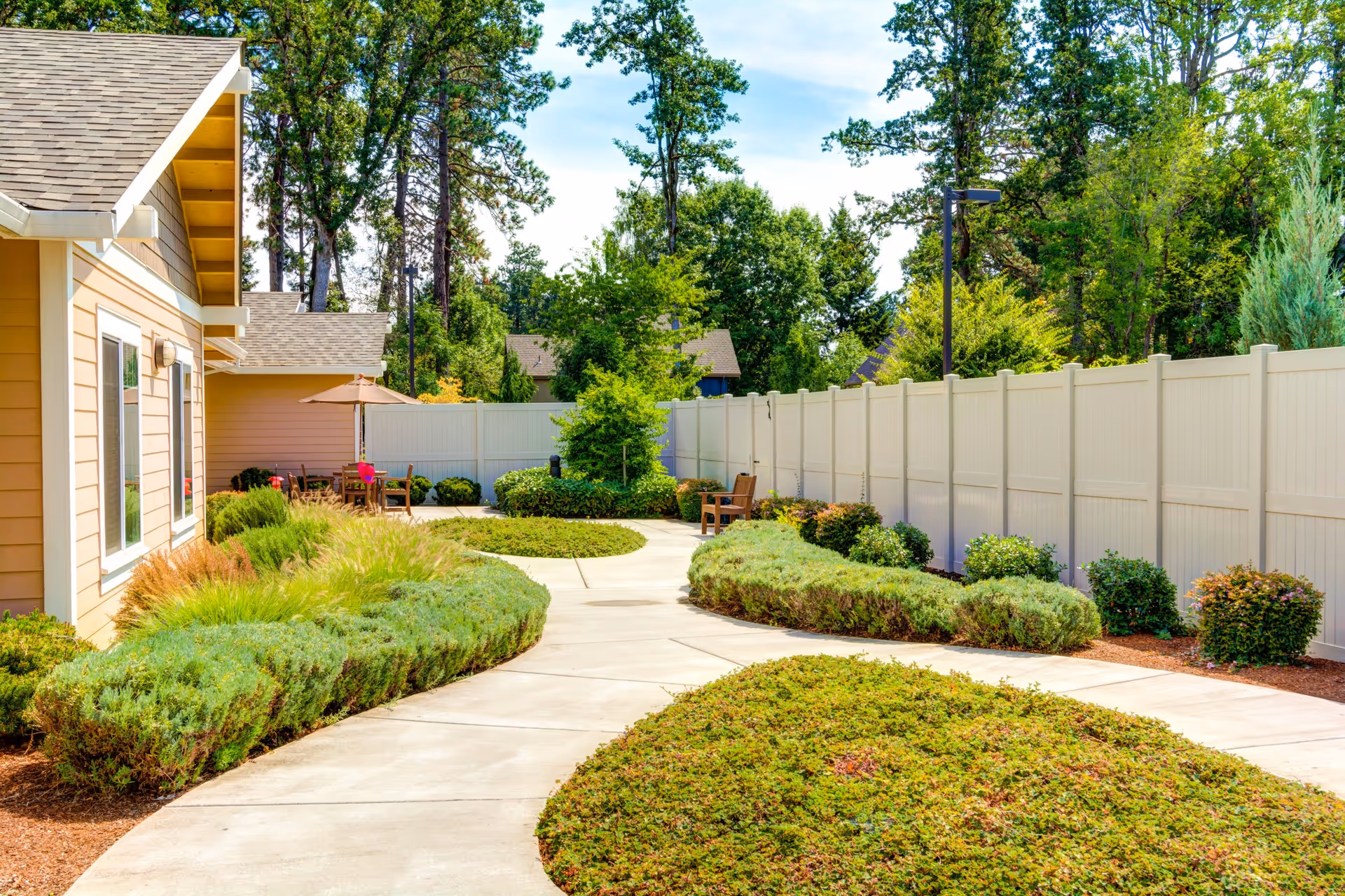 A sunny outdoor garden area at Washington Gardens Memory Care featuring a winding concrete pathway bordered by well-maintained shrubs and greenery. There are wooden benches and a table with an umbrella near the beige building with white trim. Tall trees and a white privacy fence surround the space.
