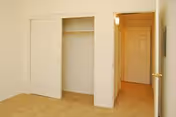 Empty bedroom with beige carpet, an open closet with sliding doors, and a hallway leading to a closed door.