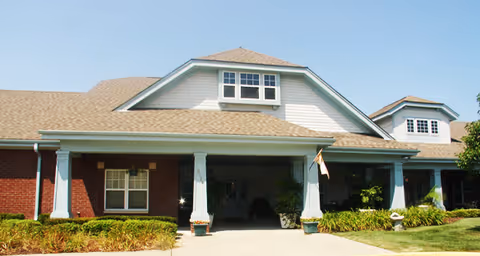 Front exterior view of a single-story building with a covered entrance supported by white columns, brick and light-colored siding, and a well-maintained lawn and shrubs under a clear blue sky.