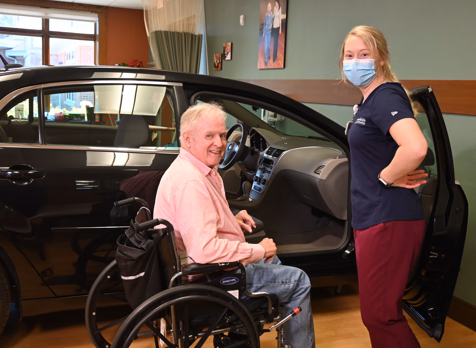 An elderly man in a wheelchair smiling next to a black car with the driver's door open inside a building. A female caregiver wearing a mask and scrubs stands nearby, also smiling. The setting appears to be indoors with green walls and framed pictures on the wall.