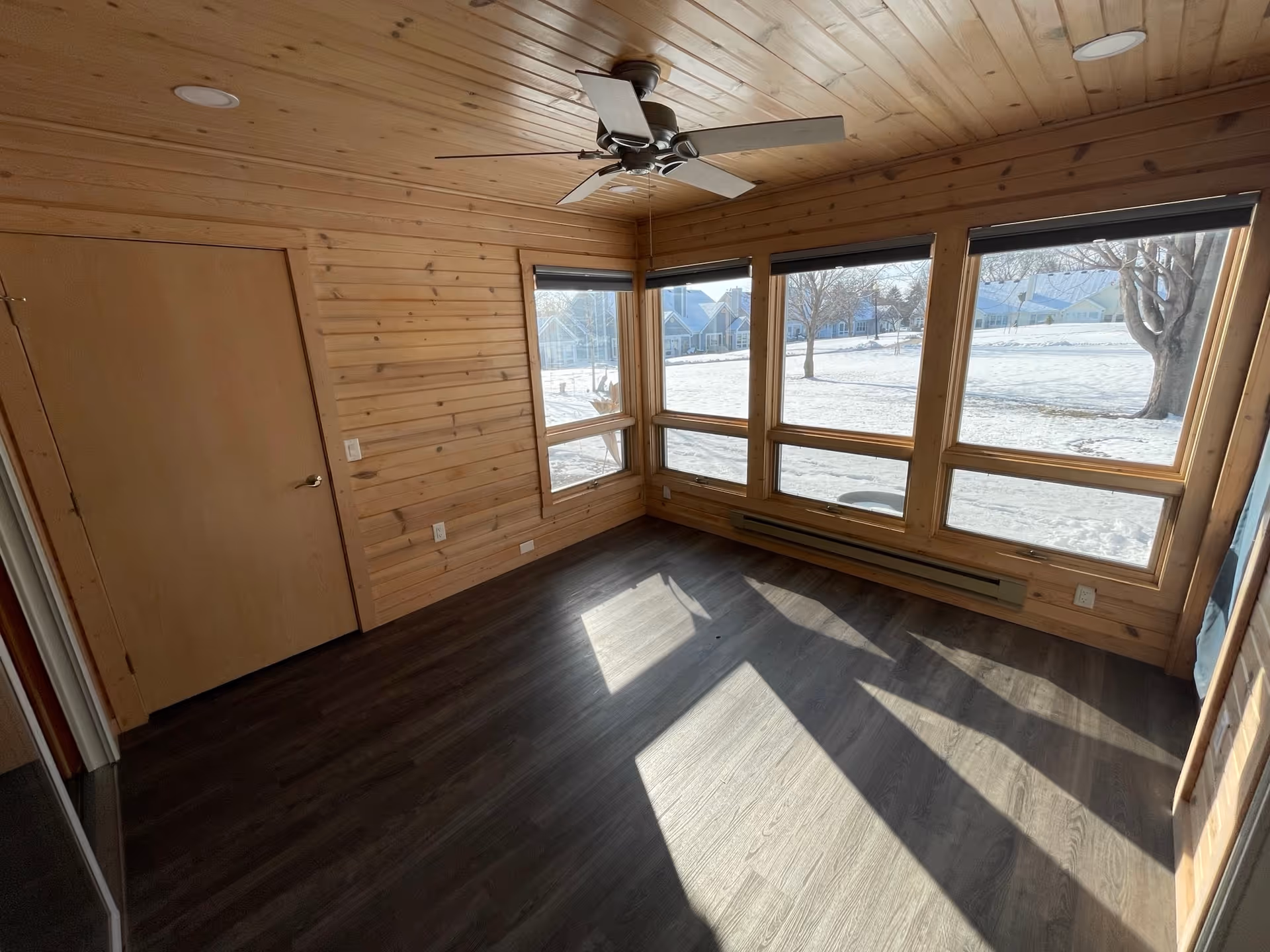 Empty room with wooden walls and ceiling, dark wood flooring, a ceiling fan, and large windows showing a snowy outdoor scene with trees and houses.