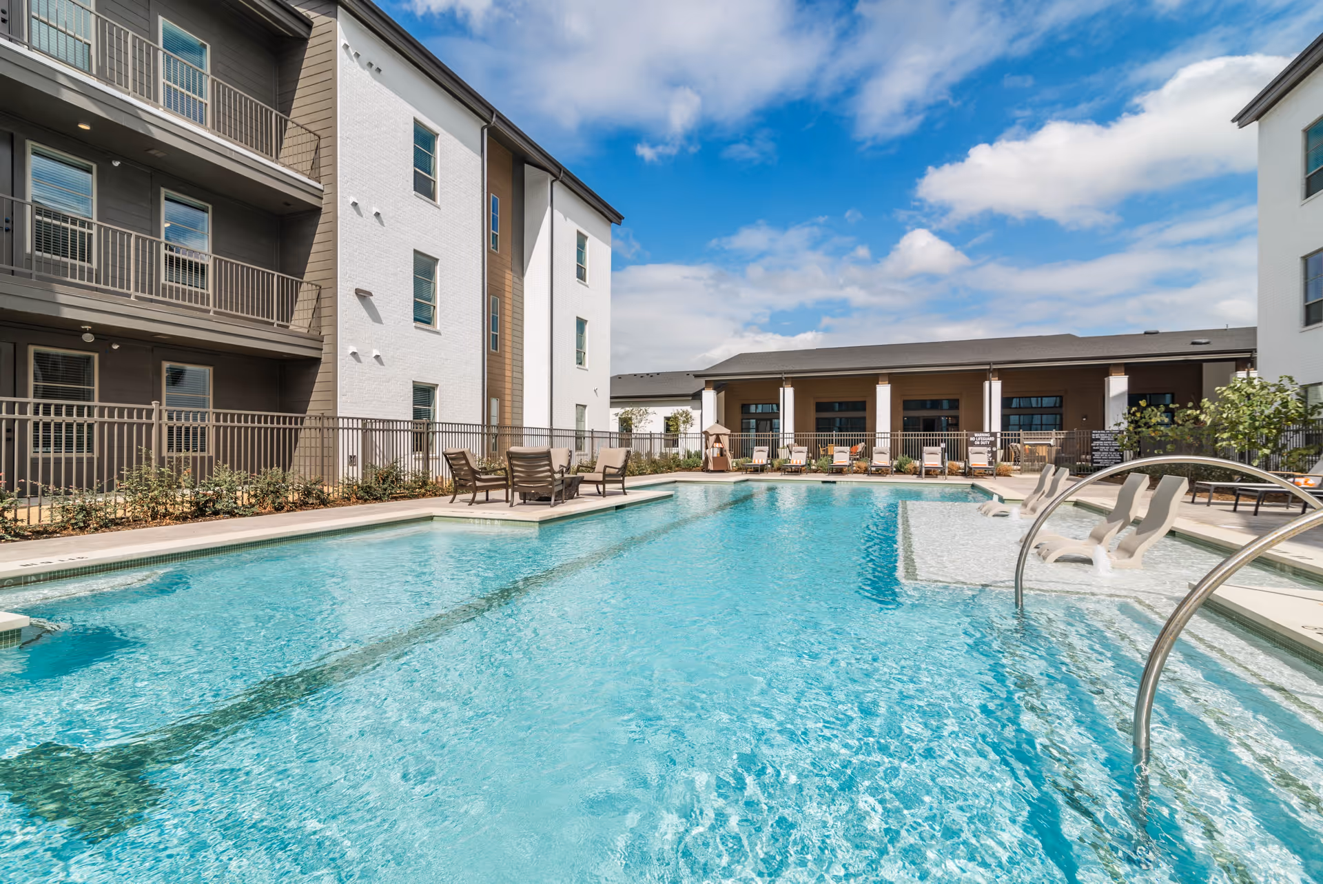 Outdoor swimming pool area at Album Benbrook with clear blue water, lounge chairs partially submerged in the pool, patio chairs and tables around the pool, and multi-story residential buildings in the background under a partly cloudy sky.