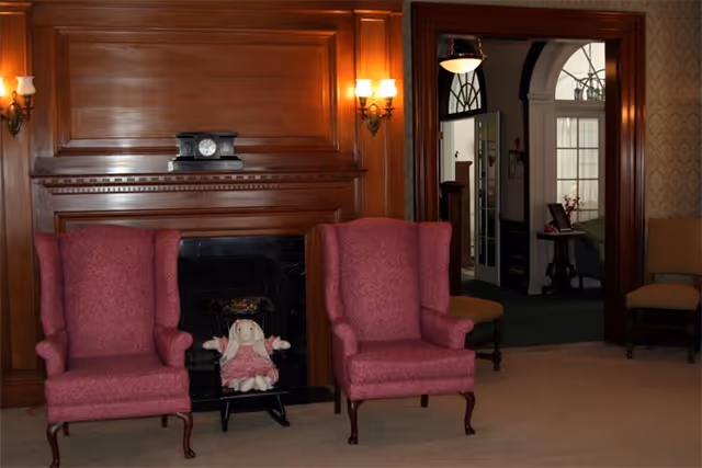 Interior seating area with two pink wingback chairs in front of a wood-paneled fireplace, a mantel clock, wall sconces, and an open doorway to another room.