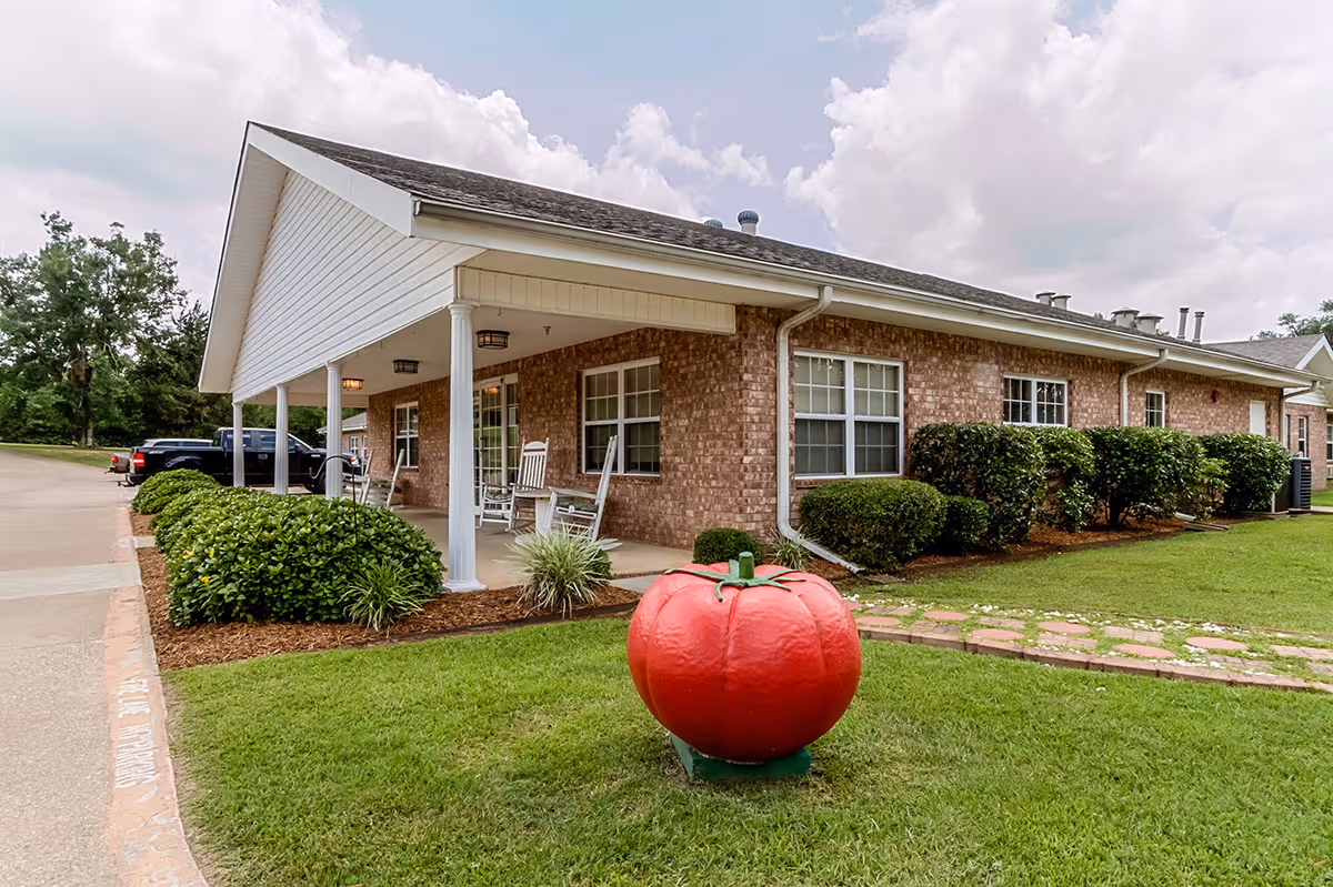 Exterior view of a single-story brick building with a covered porch featuring white rocking chairs. The building is surrounded by neatly trimmed bushes and a green lawn with a large decorative red tomato sculpture in the foreground. The sky is partly cloudy.