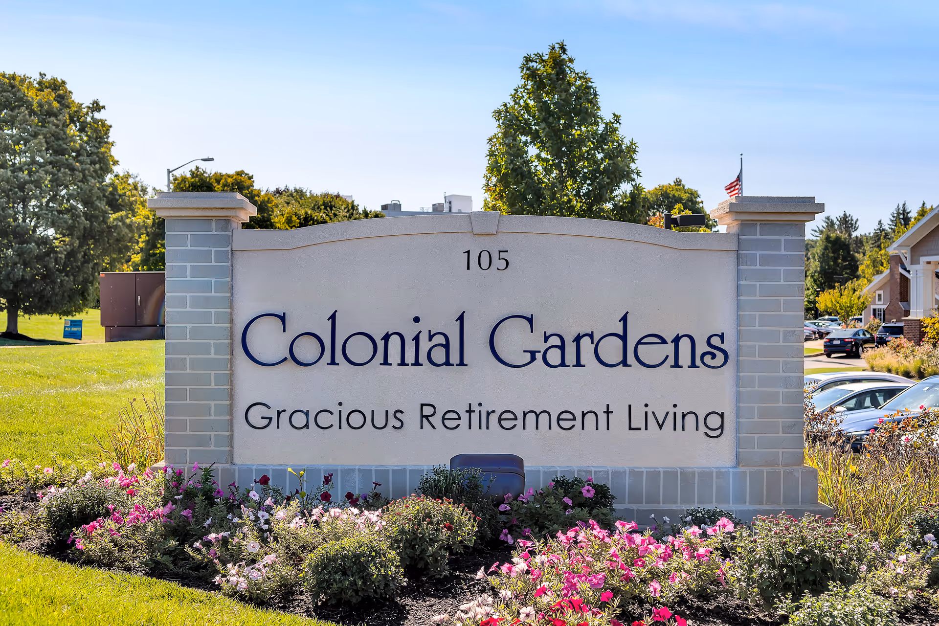 A large stone sign at the entrance of Colonial Gardens Gracious Retirement Living with the address number 105. The sign is surrounded by colorful flowers and greenery, with trees and parked cars visible in the background under a clear blue sky.