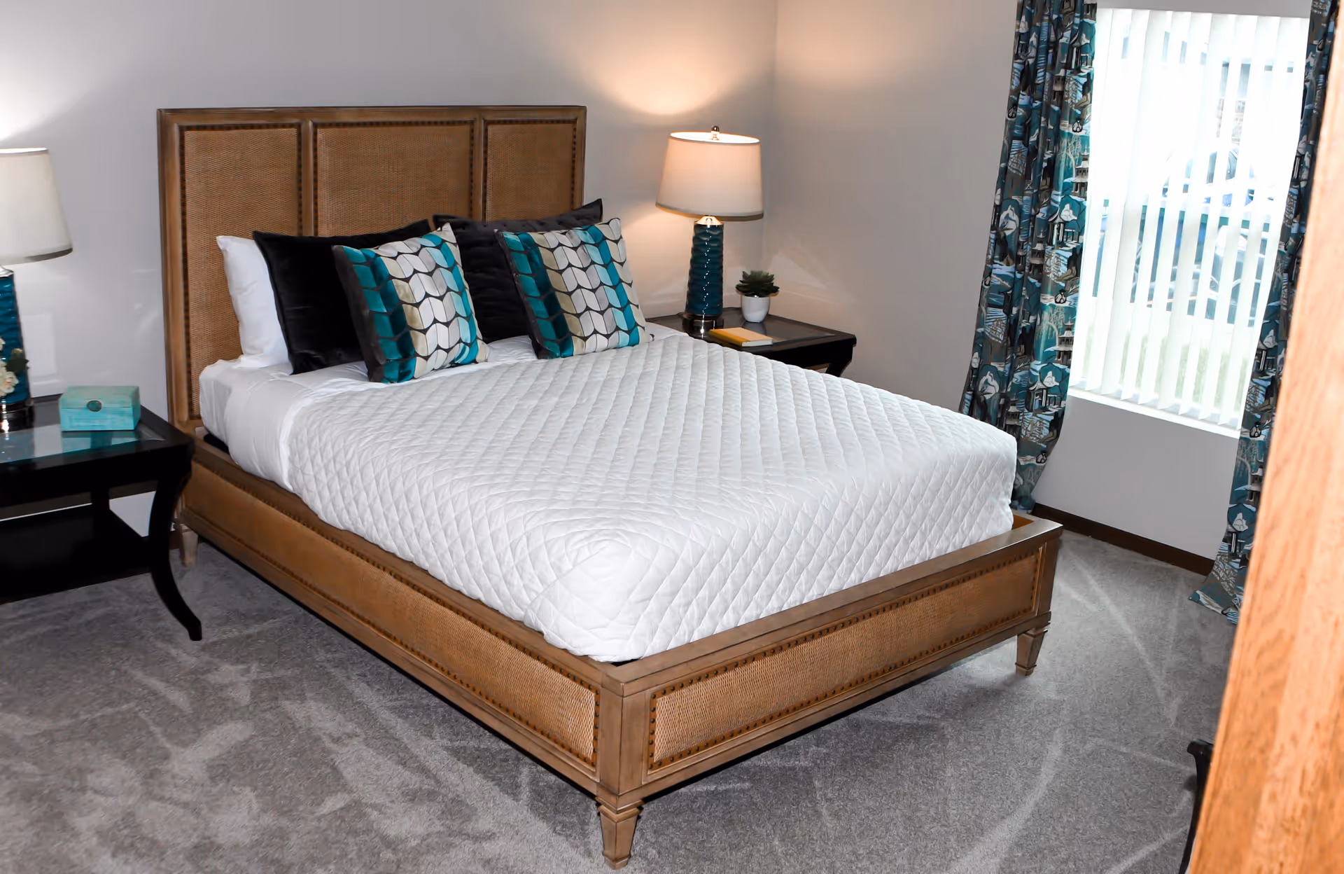 A neatly made bed with a wooden frame and a white quilted bedspread in a bedroom. The bed has four decorative pillows with teal, black, and gray patterns. There are two bedside tables with matching teal lamps and a small plant on one table. A window with vertical blinds and patterned teal curtains is visible on the right side of the room.